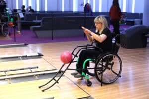 Kobieta o jasnych włosach w okularach, siedzi na wózku inwalidzkim z zielonymi akcentami. Kobieta jest ubrana na czarno. Właśnie wprowadziła różową kulę na tor za pomocą czarnej, metalowej rampy, a jej ręce są wyciągnięte. Kula stacza się z rampy w kierunku toru. W tle widoczna jest strefa wypoczynku z ciemnymi kanapami i fioletowym dywanem.