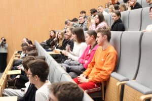 Uczestnicy wydarzenia na widowni. Młoda kobieta w białej bluzce trzyma mikrofon. Obok niej siedzą inni studenci, wszyscy patrzą w stronę prelegentów. W tle widać kolejne rzędy widowni