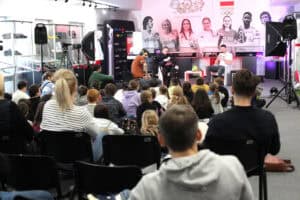 Sala z młodzieżą siedzącą na podłodze i krzesłach, w głębi Michał Szyba rozmawia z prowadzącym, pomieszczenie nowocześnie wyposażone z gablotami i oświetleniem studyjnym. Widok z widowni