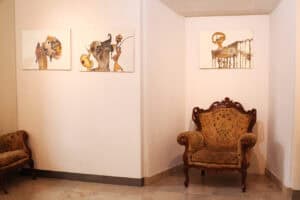 Narożnik galerii z trzema wyeksponowanymi grafikami. Po lewej stronie widać fragment fotela, a w centralnym punkcie – duży, klasyczny fotel uszak ustawiony w rogu pod jedną z prac.