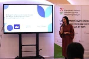 Anna Szalast trzyma mikrofon i uśmiechając się przemawia, stojąc obok baneru PFRON. Slajd dotyczy "Dialogu i koordynacji działań w obszarze lokalnego systemu wsparcia osób z niepełnosprawnościami"
