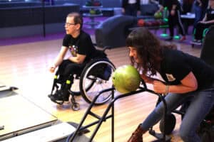 Rozgrywka w kręgle. Kobieta z ciemnymi włosami, w dżinsach i brązowych botkach, pochyla się nisko, aby rzucić zieloną kulę z metalowej rampy. Po lewej stronie, na drugim planie, mężczyzna w okularach na wózku inwalidzkim obserwuje grę, trzymając rękę w geście rzutu.