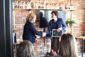 Katarzyna Herman-Gładysz i Joanna Olszewska wymieniają uścisk dłoni. Joanna Olszewska trzyma dyplom w ramce, w tle monitor i półki.