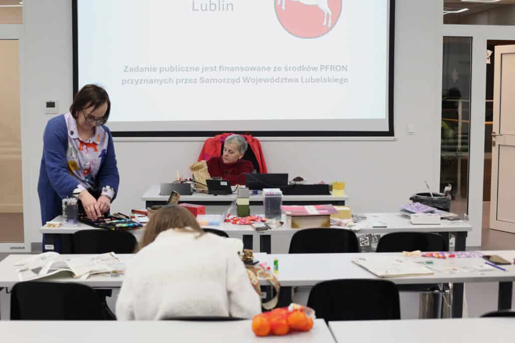 Wnętrze sali wykładowej. Prowadząca w niebieskim kardiganie (dr Anna Bieganowska-Skóra) przygotowuje przybory przy stole pod ekranem projekcyjnym. Na ekranie widnieje komunikat o finansowaniu zadania ze środków PFRON przez Samorząd Województwa Lubelskiego. Pod ekranem, za biurkiem siedzi kobieta (Krystyna Andrzejuk).