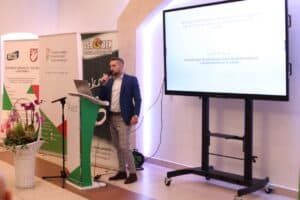 Hubert Korniszuk wygłasza prelekcję. Obok ustawiony jest ekran z wyświetloną prezentacją. Slajd dotyczy "Realizacji świadczenia wspierającego w Wojewódzkim Zespole ds. Orzekania o Niepełnosprawności w Lublinie". Z lewej stojące banery PFRON. W lewym dolnym rogu widać biały, wiklinowy kosz z ciemnoróżowymi orchideami