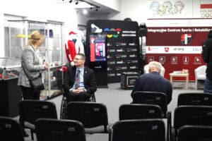 Od lewej kobieta o blond włosach, widoczna z profilu, ubrana w szary żakiet, stoi i rozmawia z mężczyzną (Piotr Jacniacki) siedzącym na wózku inwalidzkim. Mężczyzna ma na sobie garnitur i jest zwrócony do kobiety. W tle widoczny jest wystrój hali z rzędami czarnych, pustych krzeseł, witrynami wystawowymi z pucharami i odzieżą sportową oraz dużymi banerami „Integracyjne Centrum Sportu i Rehabilitacji 'Start'" oraz „Centrum Historii Sportu w Lublinie".