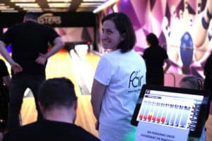 Wnętrze kręgielni. Magdalena Szpyt w białej koszulce z dużym logo 'FAR' i adresem strony internetowej na plecach. Stoi tyłem do toru do kręgli, patrzy przez ramię w kierunku aparatu i uśmiecha się. Na pierwszym planie, w prawym dolnym rogu, widoczny jest ekran tabletu wyświetlający wyniki gry w kręgle. W tle widoczny jest tor do kręgli i niewyraźne sylwetki innych graczy.