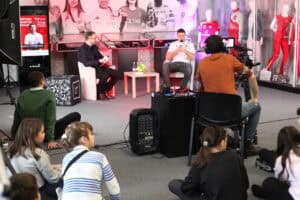 Michał Szyba siedzi na fotelu odpowiada na pytania, w dolnej części kadru dzieci siedzą na dywanie, po prawej manekiny w strojach sportowych.