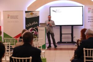 Mężczyzna w szarej marynarce w kratę przemawia ze sceny, trzyma mikrofon. Ekran za nim wyświetla logo Centrum Usług Społecznych w Świdniku. W tle widoczne są banery: PFRON i Ośrodek Szkolno-Wychowawczy. Widok z widowni