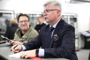 Robert Litwiński w eleganckim, granatowym garniturze w kratę i okularach. Mężczyzna trzyma długopis nad otwartą książką i patrzy w bok. Na jego nadgarstku widać smartwatch. W tle, z lewej strony, stoi inny, uśmiechnięty mężczyzna w okularach i oliwkowej kurtce, trzyma w ręku broszury - jego sylwetka jest nieostra.