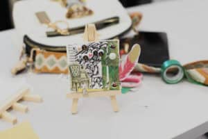 Ukończona praca plastyczna umieszczona na małej, drewnianej sztaludze stojącej na białym stole. Kolaż zawiera imię „KAMIL" ułożone z koralików z literami, grafikę fotela, piórko oraz kolorowe kamyczki ozdobne. W tle widać białą torebkę i akcesoria warsztatowe.