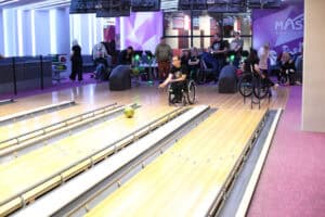 Wnętrze nowoczesnej kręgielni „MASTERS BOWLING" z fioletowym dywanem i fioletowo-białymi ścianami. Mężczyzna w okularach, na wózku inwalidzkim w czarnym T-shircie, rzuca zieloną kulę, która toczy się na jasnym, drewnianym torze. W tle widoczna jest grupa ludzi siedzących i stojących, z których niektórzy również są na wózkach.