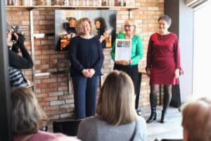 Katarzyna Herman-Gładysz, Magdalena Krawczyk oraz uczestniczka w zielonym swetrze pozują z certyfikatem przed ceglaną ścianą. Z boku widoczna osoba fotografująca.