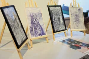 Cztery małe grafiki w ramkach stoją na drewnianych sztalugach. Na blacie leży plakat wystawy Jana Kaweckiego.