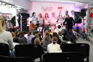 Prowadzący podchodzi do dziecka na podłodze, aby mogło zadać pytanie, Michał Szyba obserwuje z fotela, po bokach lampy studyjne i statywy z kamerami, w tle zdjęcia olimpijczyków i trofea. Widok z widowni