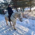 Agnieszka Kwolek siedzi na białym koniu Kira w zimowej scenerii na terenie Fundacji Horses and Family. Kobieta ma zimową odzież i kask jeździecki; fot. Barbara Jarząb