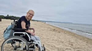 Na piaszczystej plaży siedzi w wózku uśmiechnięta Aleksandra. Ma blond włosy związane w kucyk, okulary w czarnej oprawie i szeroko się uśmiecha. W tle widać panoramę morza, spienione fale przy brzegu i zieleń w oddali.