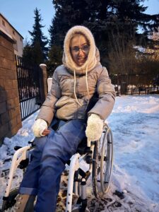 Przed ogrodzeniem na zewnątrz w zimowej aurze siedzi uśmiechnięta dziewczyna w okularach, ubrana w zimową odzież. Wokół leży śnieg, niebo jest błękitne, a na drzewa w tle świeci późnopopołudniowe słońce