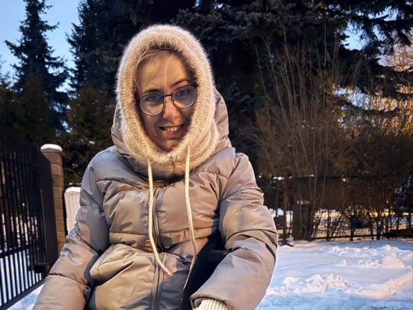 Na zewnątrz w zimowej aurze siedzi uśmiechnięta Aleksandra Górska (autorka artykułu) w okularach, ubrana w beżową zimową kurtkę i włochaty, wełniany kaptur