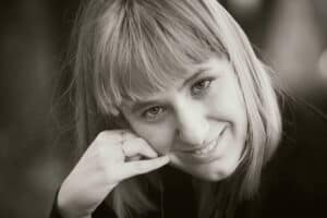 Czarno-biały portret kobiety z grzywką. Podpiera policzek prawą dłonią, na palcach ma obrączkę na serdecznym- i pierścionek z niewielkim oczkiem na środkowym palcu. Uśmiecha się i patrzy wprost w kadr; fot. Grzegorz Kominek – NPICTURES