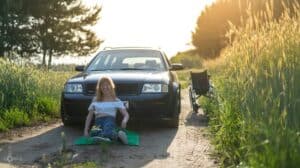 Na wiejskiej, piaszczystej drodze pomiędzy zielonymi polami zbóż stoi czarny samochód marki Audi. Obok auta stoi wózek inwalidzki. Przed przednią maską samochodu na zielonym kocu siedzi blondynka w okularach, ubrana w białą bluzkę typu ‘hiszpanka’ i niebieskie jeansy. Kobieta szeroko się uśmiecha. Słońce rozświetla czubek jej głowy i włosy, choć większość ciała znajduje się w cieniu samochodu; fot. Paweł Pietrzak - Hokus Pokus