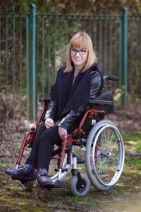 Ocieniona ścieżka z mchem i zieloną trawką w plenerze, z prześwitami słońca i zielonym ogrodzeniem w tle. Na ścieżce na wózku siedzi uśmiechnięta kobieta w okularach z czarną oprawką, z włosami obciętymi na pazia i grzywką; fot. Grzegorz Kominek – NPICTURES