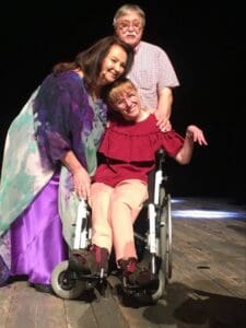 Na scenie podczas Ogólnopolskiego Festiwalu Twórczości Teatralno-Muzycznej Osób Niepełnosprawnych "Albertiana 2022" Anna Dymna lekko pochyla się i przytula siedzącą na wózku Aleksandrę Górską. Za wózkiem stoi instruktor muzyczny Marek Kowal i trzyma lewą dłoń na ramieniu Aleksandry