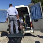 Ateny, Epidaurus, 2017. Bus z opuszczonym podnośnikiem. Na platformie stoi wózek, za nim mężczyzna. W środku pojazdu znajduje się drugi mężczyzna, który oczekuje na pasażerkę. Słoneczna pogoda, błękitne niebo