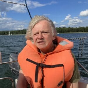 Mężczyzna w pomarańczowej kamizelce płynie łodzią. W oddali widać białe łódki i żaglówki, a na horyzoncie las. Wiatr porusza włosy mężczyzny