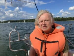 Sierpień 2025, Jezioro Kisajno, Ośrodek Oczy Mazur. Mężczyzna w pomarańczowej kamizelce płynie łodzią. W oddali widać białe łódki i żaglówki, a na horyzoncie las. Wiatr porusza włosy mężczyzny