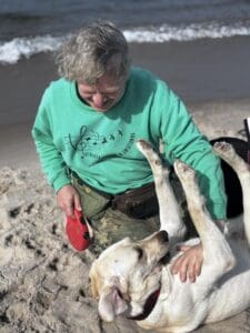 Kwiecień 2025, Jastrzębia Góra, Władysławowo. Na plaży nad brzegiem morza mężczyzna w zielonej bluzie z napisem ‘Otwarta Przestrzeń Światłownia’ bawi się z psem przewodnikiem – kremowym labradorem Ramenem. Pies leży na plecach i unosi przednie łapy, a mężczyzna trzyma dłoń na jego klatce piersiowej