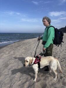 Kwiecień 2025, Jastrzębia Góra, Władysławowo. Na plaży nad morzem stoi mężczyzna w zielonej bluzie z napisem ‘Otwarta Przestrzeń Światłownia’, na plecach ma czarny plecak, trzyma białą laskę i prowadzi psa przewodnika – kremowego labradora Ramena