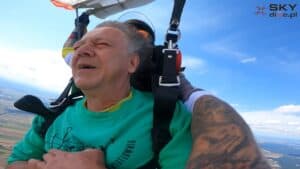 Lipiec 2024, Lotnisko Włocławek-Kruszyn. Mężczyzna w zielonej bluzie z napisem ‘Otwarta Przestrzeń Światłownia’ skacze ze spadochronem w tandemie. Ma zamknięte oczy, włosy zaczesane do tyłu przez wiatr, jest w uprzęży z widocznymi szelkami. W tle błękitne niebo z białymi obłoczkami i ziemia widziana z dużej wysokości