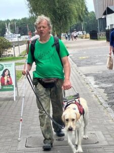 Mężczyzna z psem przewodnikiem idzie chodnikiem. Ma zieloną koszulkę, spodnie moro, czarny plecak i saszetkę. Trzyma białą laskę, psa prowadzi ręką. W tle ludzie, drzewa, jachty i fragment budynku, po lewej stoi potykacz reklamowy z kobietą i napisem o lodach