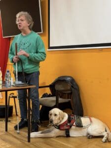 Październik 2025, Ośrodek Szkolno-Wychowawczy w Bydgoszczy. Grzegorz Dudziński prowadzi zajęcia ‘Instrukcja Obsługi Niewidomego’. W sali o żółtych ścianach stoi mężczyzna w zielonej bluzie z napisem ‘Otwarta Przestrzeń Światłownia’, trzyma białą laskę i gestykuluje. Obok jego lewej nogi leży kremowy pies przewodnik w uprzęży.