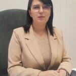 Portret biznesowy. W jasnym wnętrzu przy biurku siedzi brunetka w okularach, ubrana w beżową bluzkę i żakiet. Dłonie ma ułożone na zamkniętym laptopie i patrzy w obiektyw z łagodnym wyrazem twarzy