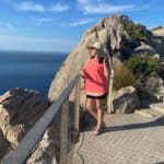 Słoneczny taras nad morzem na skałach. Przy barierce stoi kobieta z długimi ciemnymi włosami w letnim stroju, kapeluszu i okularach przeciwsłonecznych. Lewą ręką przytrzymuje kapelusz i patrzy w stronę horyzontu.