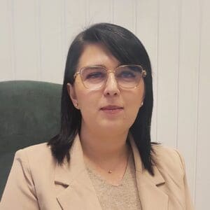 Portret biznesowy. W jasnym wnętrzu przy biurku siedzi brunetka w okularach, ubrana w beżową bluzkę i żakiet.