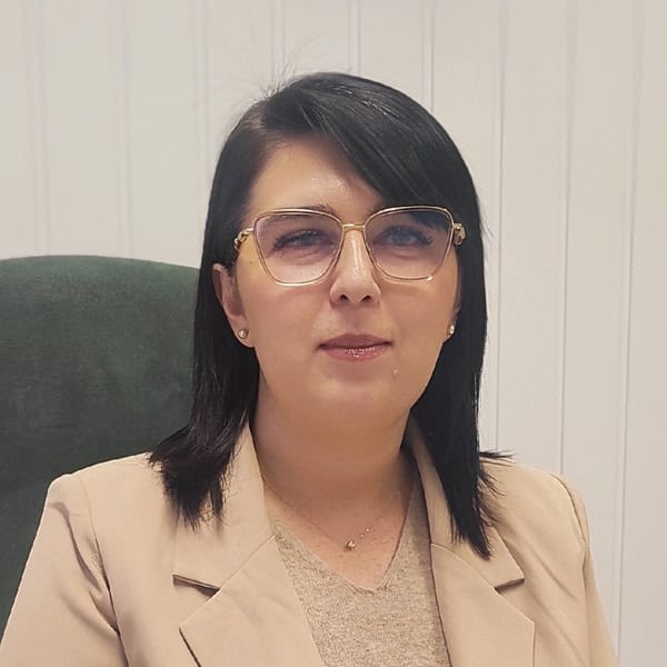 Portret biznesowy. W jasnym wnętrzu przy biurku siedzi brunetka w okularach, ubrana w beżową bluzkę i żakiet.