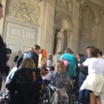 Paryż, 2019. Turyści zwiedzają muzealne wnętrze z płaskorzeźbami, rzeźbą i zdobionymi drzwiami, podłoga w biało-czarne płytki. W tłumie znajdują się osoby na wózkach