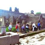 Pompeje, 2024. Grupa ośmiu osób porusza się jeden za drugim pomiędzy murami starożytnych zabudowań ulicznych. W grupie są osoby na wózkach z opiekunami