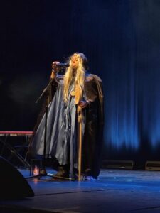 Występ wokalny Sebastiana podczas 28. Spotkań z Piosenką Nieprzetartego Szlaku „Fantasy” (27–29 października 2025). Mężczyzna w stroju maga ma na sobie długą czarną pelerynę z kapturem i perukę z długimi blond włosami. Prawą ręką trzyma mikrofon na statywie, w lewej ręce kostur; fot. Anna Bieganowska-Skóra