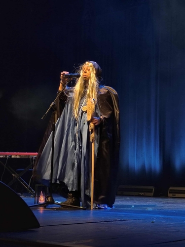 Występ wokalny Sebastiana podczas 28. Spotkań z Piosenką Nieprzetartego Szlaku „Fantasy” (27–29 października 2025). Mężczyzna w stroju maga ma na sobie długą czarną pelerynę z kapturem i perukę z długimi blond włosami. Prawą ręką trzyma mikrofon na statywie, w lewej ręce kostur; fot. Anna Bieganowska-Skóra