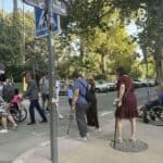 Sewilla, Hiszpania, 2025. Przez przejście dla pieszych przechodzi grupa osób, w tym osoby z niepełnosprawnością ruchową o kulach i laskach, osoby na wózkach pchanych przez asystentów oraz osoby sprawne