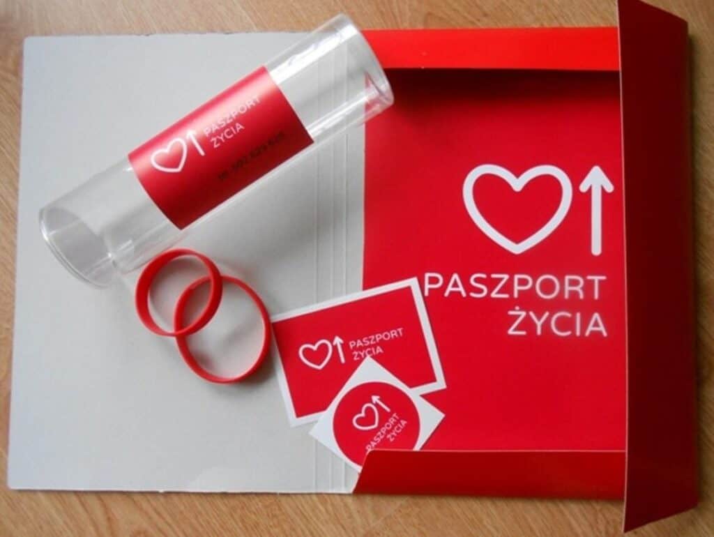 Na blacie leżą materiały z logo i oznaczeniami "Paszport Życia": teczka, opaski, naklejki i cylindryczne etui
