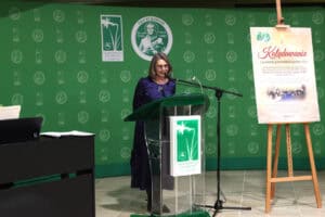 prof. Iwona Kalinowska w ciemnoniebieskiej sukience przemawia przy szklanej mównicy na tle zielonej ścianki z logo Centrum Onkologii Ziemi Lubelskiej im. św. Jana z Dukli. Po prawej stronie stoi sztaluga z plakatem zapraszającym na wspólne kolędowanie, a po lewej fragment czarnego instrumentu klawiszowego.