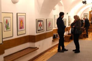 Kobieta i mężczyzna rozmawiają w jasnym pomieszczeniu galerii o łukowym sklepieniu. Na białych ścianach wiszą oprawione grafiki przedstawiające kolorowe kwiaty, a pod nimi ciągną się drewniane ławy. W tle widać wejście do kolejnej sali i inne osoby uczestniczące w wydarzeniu.