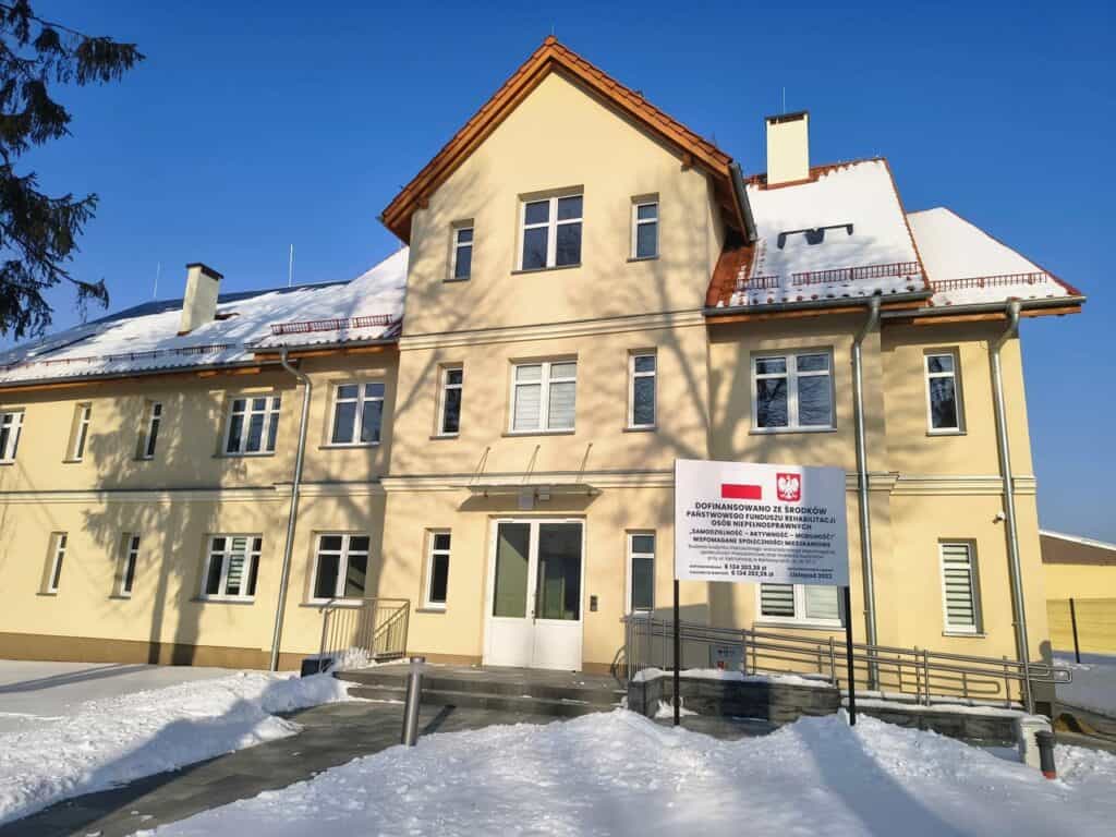 Piętrowy budynek Wspomaganej Społeczności Mieszkaniowej w Bartoszycach w zimowej scenerii, z pokrytym śniegiem otoczeniem i błękitnym niebem w tle