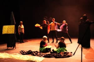 Zbiorowa scena teatralna na ciemnej scenie. Na środku dwie osoby w czarnych bluzkach i zielonych spódnicach siedzą na podłodze. Za nimi stoi grupa aktorów: mężczyzna trzymający suche gałęzie oraz osoby z długimi pasami pomarańczowego i fioletowego tiulu. Po prawej stronie widać dwie postacie w długich, czarnych habitach z zakrytymi twarzami. Po lewej stoi kobieta w szarym t-shircie, a obok niej sztaluga przykryta kolorowym materiałem w kwiaty. Całość oświetlona jest ciepłym, punktowym światłem