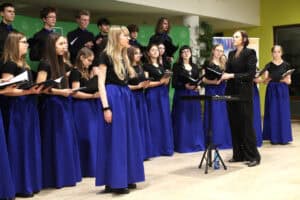 Zbliżenie na występ chóru młodzieżowego. Na pierwszym planie młoda kobieta o blond włosach, w okularach śpiewa solo, ubrana w czarną koszulkę i długą chabrową spódnicę. Obok niej, po prawej stronie, stoi dyrygentka w czarnym, połyskującym garniturze, patrząc w stronę zespołu. W tle pozostali członkowie chóru z nutami w dłoniach oraz biały baner informacyjny.